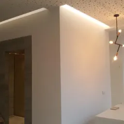 Białe ściany po malowaniu z podświetlanym sufitem i nowoczesną lampą wiszącą, widoczny fragment łazienki z umywalką i dozownikiem mydła.