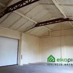 Wnętrze magazynu po natryskowej izolacji pianką PUR, widoczne elementy konstrukcji stalowej i brama wjazdowa, logo firmy w prawym dolnym rogu.