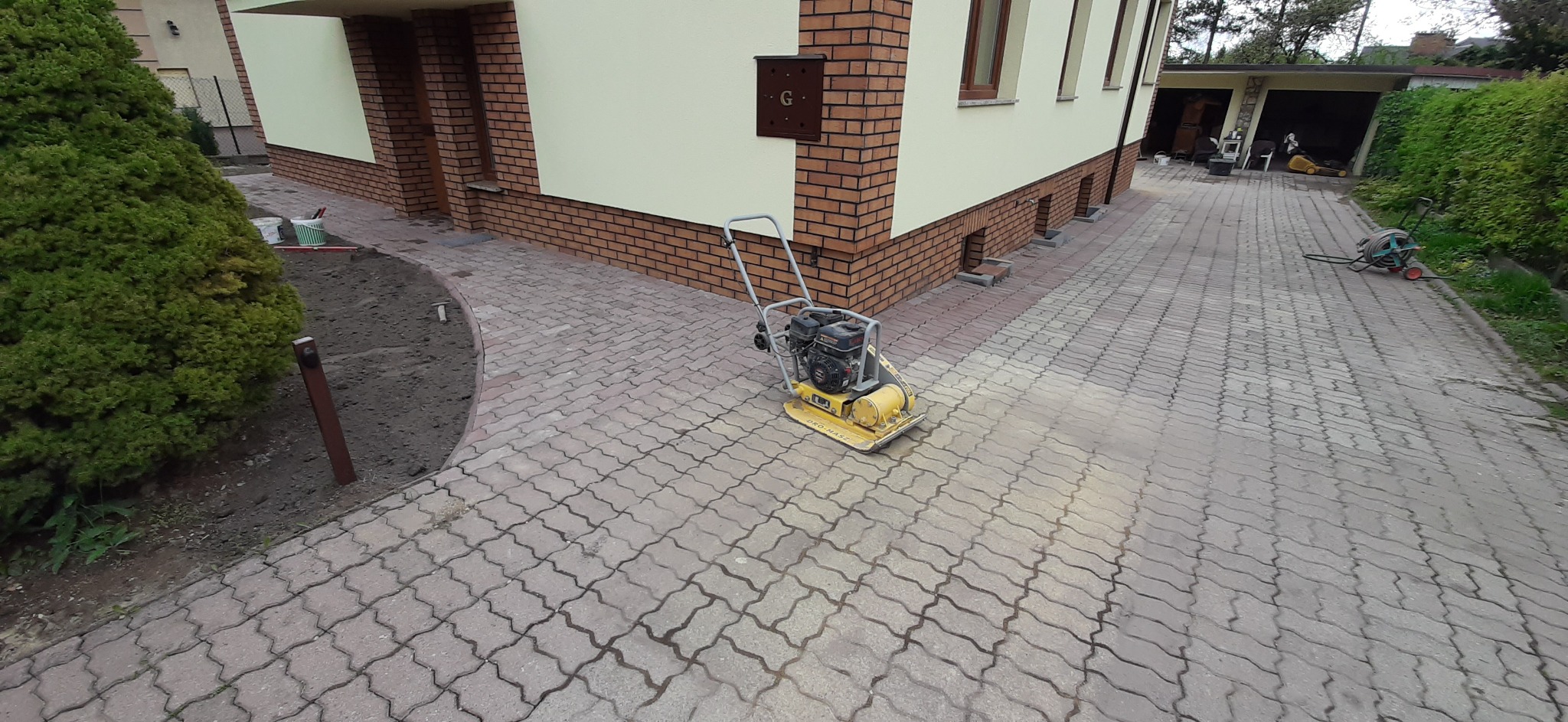 Żółta zagęszczarka na ułożonej kostce brukowej przed budynkiem z ceglaną podmurówką i przygotowanym miejscem na rabatę