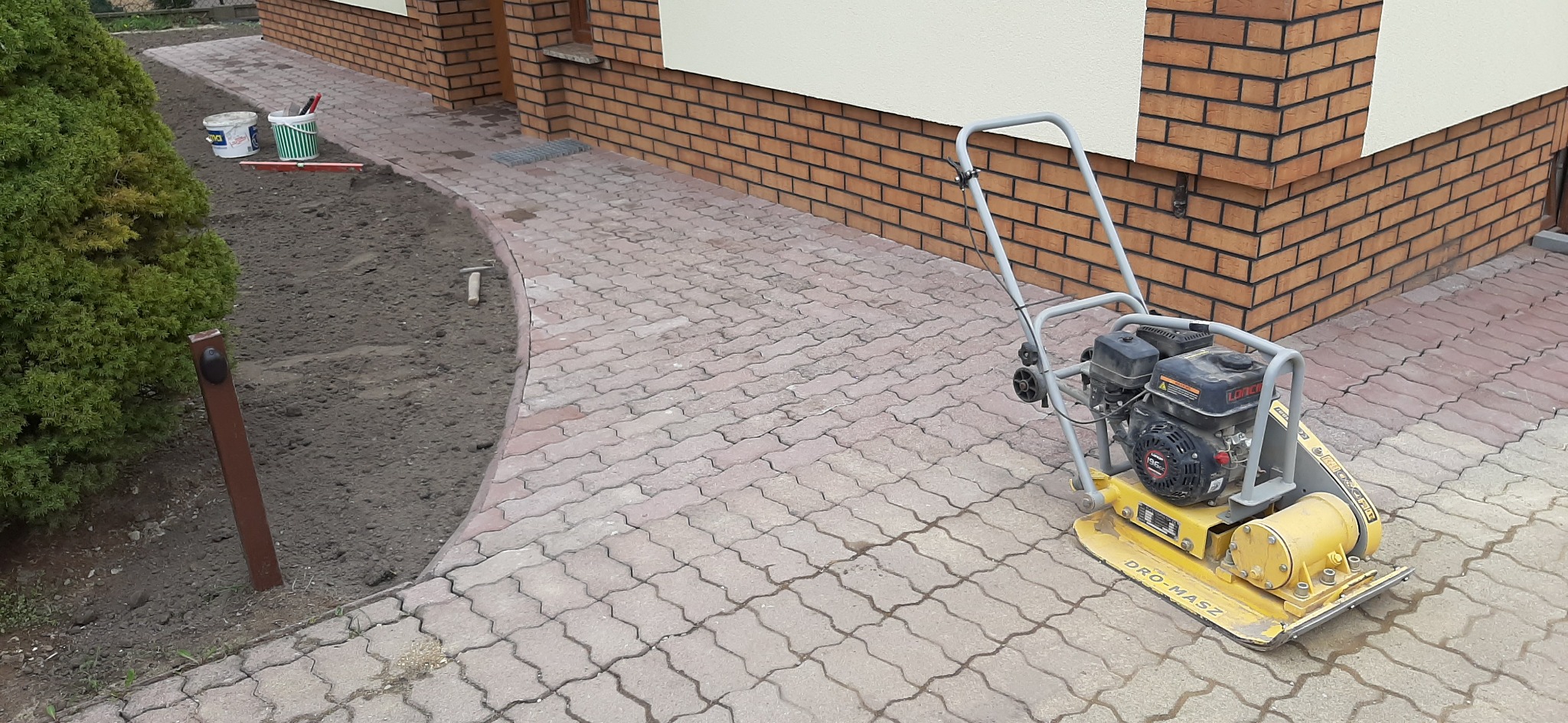 Żółta zagęszczarka spalinowa na ułożonej kostce brukowej, obok przygotowany teren pod trawnik, w tle ściana budynku z cegły klinkierowej.