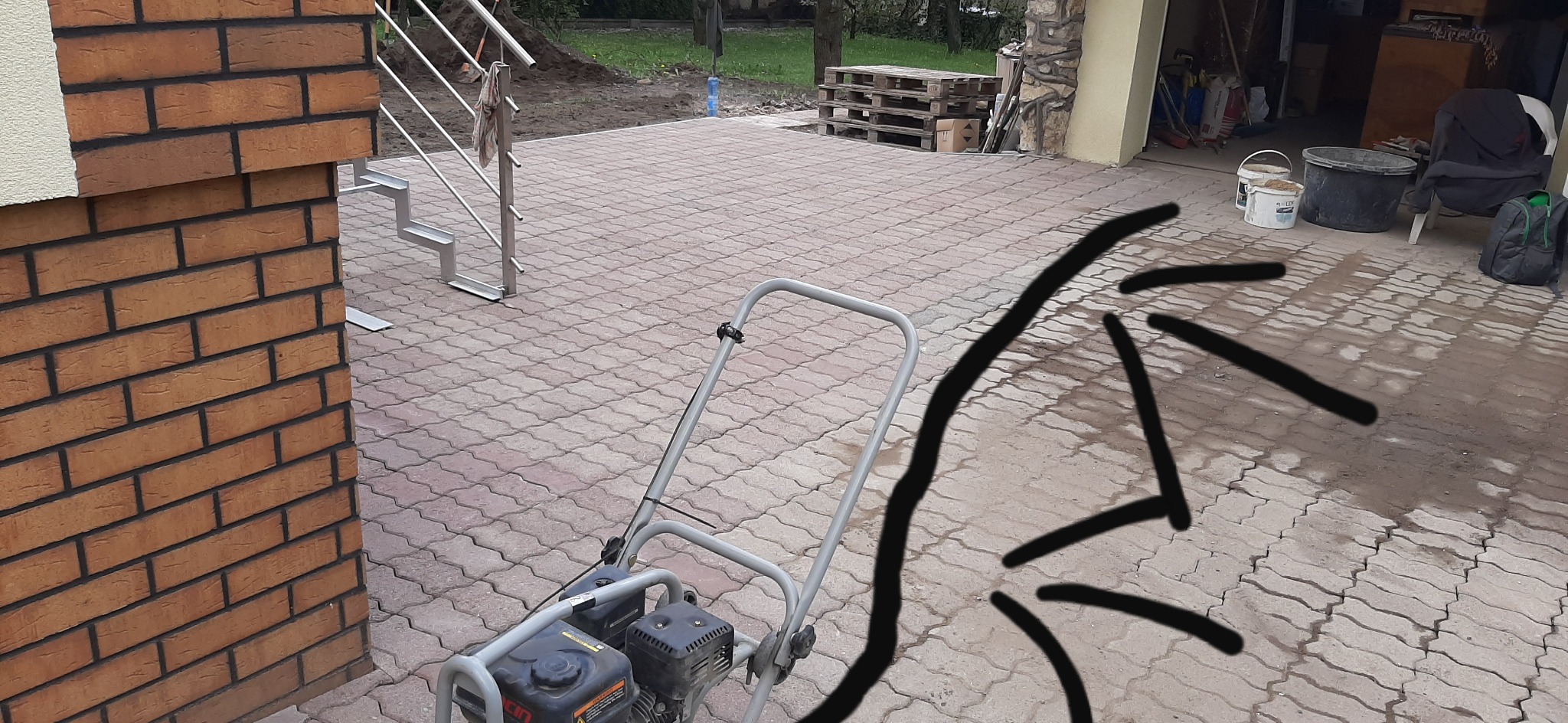 Wibrująca zagęszczarka stoi na nowo ułożonej kostce brukowej w kształcie fali, obok ceglanego narożnika budynku i metalowych schodów zewnętrznych.