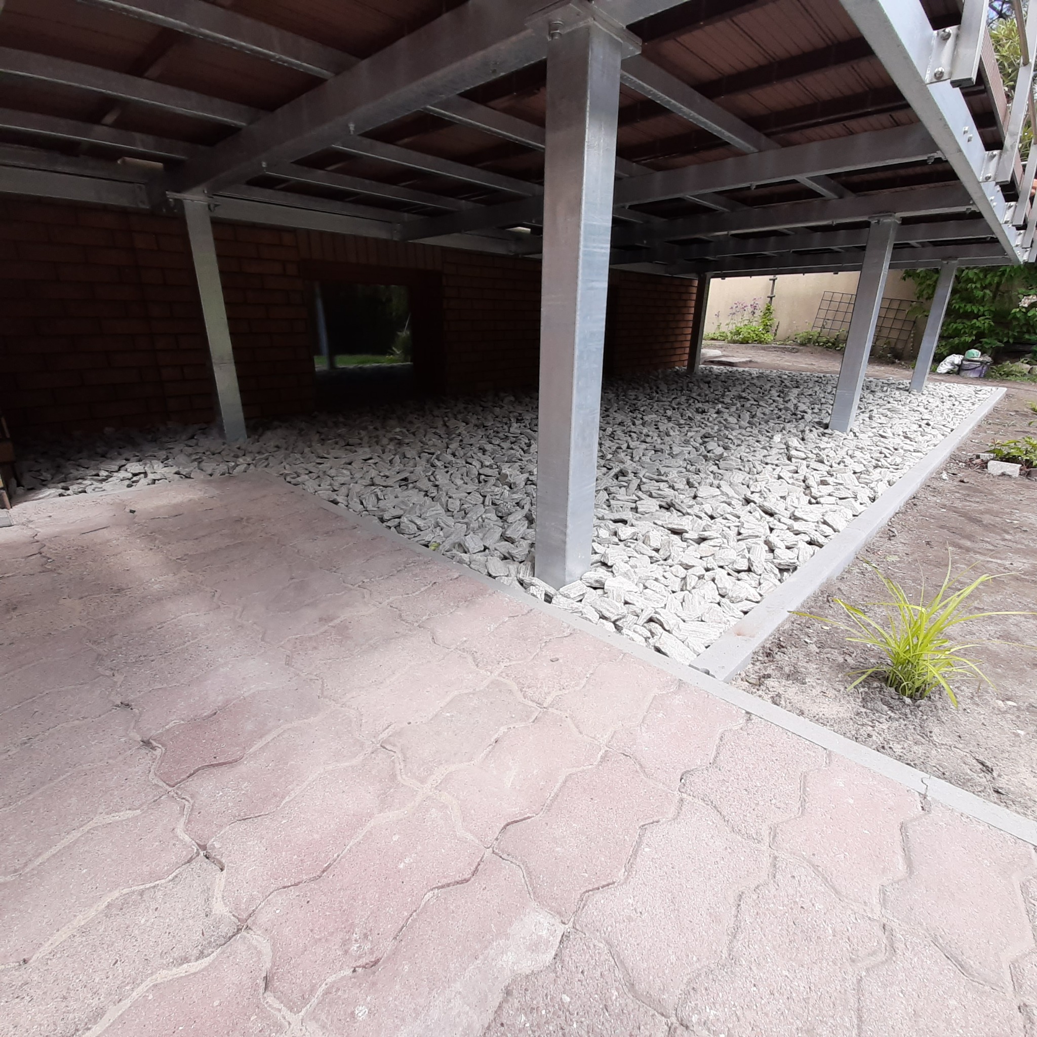 Zadaszenie tarasu z metalową konstrukcją, podsypane białym kamieniem, z fragmentem różowej kostki brukowej na pierwszym planie i ceglaną ścianą w tle.