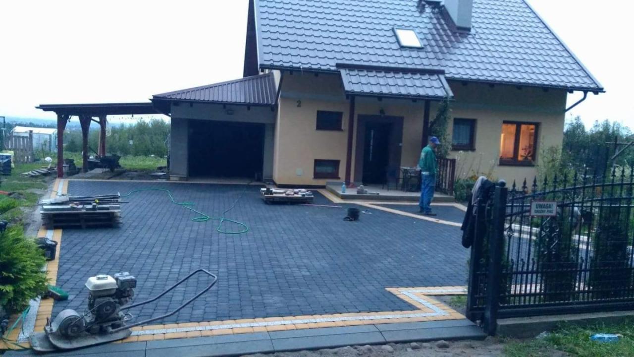 Ułożona kostka brukowa w kolorze grafitowym z żółtymi obrzeżami przed domem jednorodzinnym z garażem, widoczna zagęszczarka wibracyjna do ubijania kostki, paleta z materiałami i pracownik w oddali.