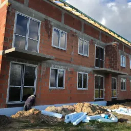 Wznoszony dwukondygnacyjny budynek z czerwonej cegły z zamontowanymi oknami; widoczna warstwa izolacji fundamentów i pracownik przy dolnym oknie.