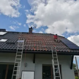 Pracownik w czerwonej koszulce siedzi na dachu z dachówki ceramicznej, montując panele fotowoltaiczne na aluminiowej konstrukcji, widoczne dwie drabiny opierające się o ścianę budynku, błękitne...