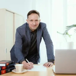 Uśmiechnięty mężczyzna w garniturze pochyla się nad biurkiem, pisząc na kartce; obok laptop, filiżanka kawy i stos książek z czerwonymi grzbietami, za nim jasne biuro z roślinami doniczkowymi.