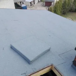 Szary, płaski dach pokryty papą termozgrzewalną z widocznym kominem z cegły i wyłazem dachowym. W tle budynki gospodarcze i drzewa.