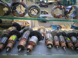 Różne wirniki i stojany elektronarzędzi, ułożone rzędami na półce w serwisie. Widoczne uzwojenia miedziane i elementy przekładni. Części zamienne do elektronarzędzi.