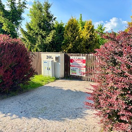 Ogłoszenie serwisu elektronarzędzi na brązowej bramie posesji z numerem 244A, otoczonej zielenią i krzewami. Widok z drogi dojazdowej.