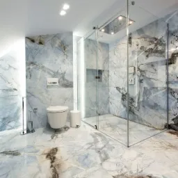 Nowoczesna łazienka z marmurowymi płytkami w odcieniach błękitu i szarości, kabina prysznicowa ze szklanymi ścianami i deszczownicą, podwieszana toaleta i minimalistyczne akcesoria.
