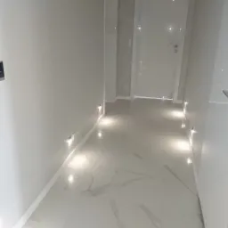 Nowoczesny korytarz z podświetlaną podłogą z płytek imitujących marmur, panelem sterowania na ścianie i tapetą w ornamenty.