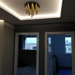 Nowoczesny sufit z podświetleniem LED i złotą lampą wiszącą z rurek, widok z dołu na szare ściany i fragmenty widoku przez dwa otwarte drzwi.