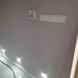 Szara ściana z zamontowanym panelem sterowania alarmem, czterema włącznikami światła i punktowym oświetleniem podłogowym, widoczny kabel, listwa przypodłogowa i fragment drewnianej framugi drzwi.