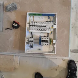 Otwarta, biała skrzynka rozdzielcza z zainstalowanymi bezpiecznikami i przewodami, leżąca na podłodze w trakcie montażu, obok miara zwijana, poziomica i fragment buta na tle surowej podłogi.