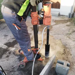 Pracownik w odzieży roboczej używa wiertnicy rdzeniowej do wiercenia w betonowej powierzchni, widoczne mokre podłoże i elementy ogrodzenia w tle.