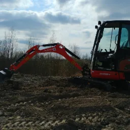 Pomarańczowa minikoparka Kubota KX018-4 na nierównym terenie z widocznymi śladami gąsienic, w tle drzewa i pochmurne niebo.