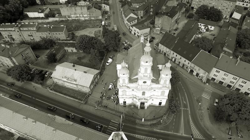 Czarno-białe ujęcie z lotu ptaka na barokowy kościół z wieżami, otoczony zabudową miejską i ulicami, z grupą ludzi przed wejściem.