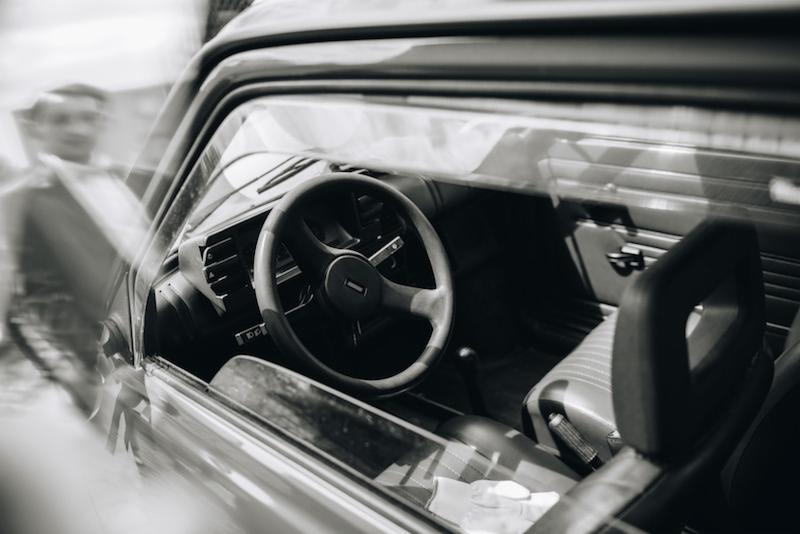 Czarno-białe zdjęcie wnętrza klasycznego samochodu, widoczne przez szybę, z ostrością na kierownicę i deskę rozdzielczą, z rozmytą postacią w tle.