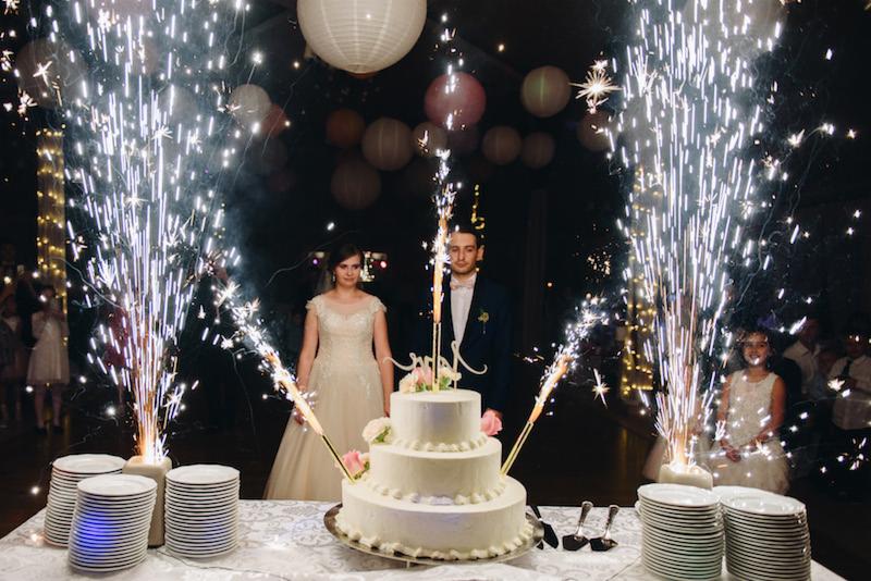 Młoda para stoi za trzy piętrowym tortem weselnym, odpalone zimne ognie tworzą efektowną oprawę podczas uroczystości.