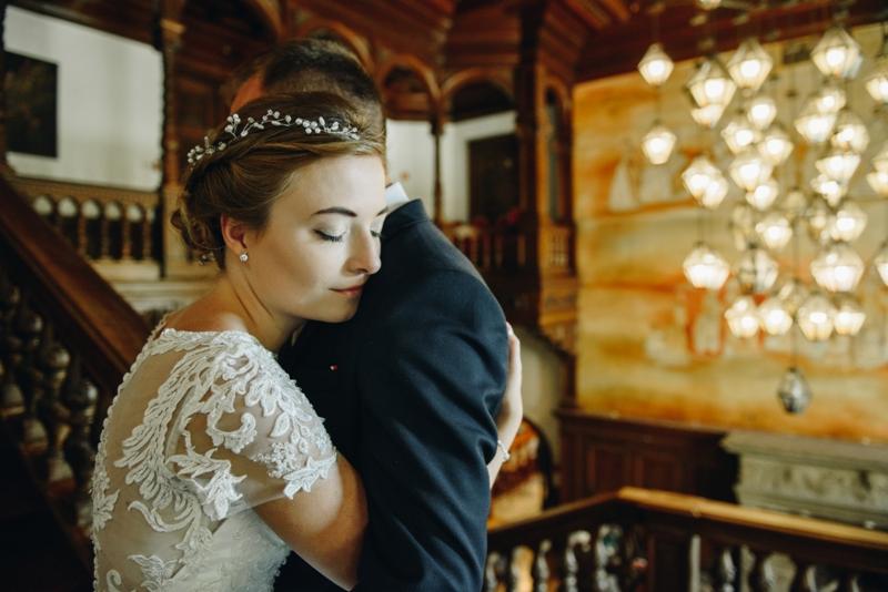 Panna młoda w koronkowej sukni ślubnej, z zamkniętymi oczami, obejmuje mężczyznę w ciemnym garniturze na tle bogato zdobionych schodów i wiszących lamp.