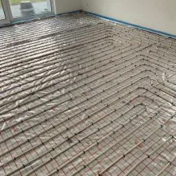 Rozłożone rury ogrzewania podłogowego na folii z czerwoną siatką w jasnym pomieszczeniu, zabezpieczone niebieską taśmą przy ścianach.