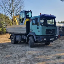 Ciężarówka MAN 19-403 z żółtą mini koparką Wacker Neuson na platformie załadunkowej, teren budowy w tle, drzewa bez liści, słoneczny dzień.