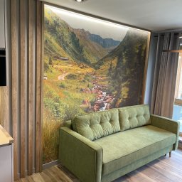 Vitalii Savchenko - Salon z zieloną sofą na tle fototapety przedstawiającej górski krajobraz z potokiem, oświetlonej listwą LED, obok drewniane lamele na ścianie i szare zasłony przy oknie.
