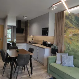 Nowoczesna kuchnia z szarymi szafkami, drewnianym blatem, stołem z czarnymi krzesłami i zieloną sofą na tle fototapety z górskim krajobrazem.