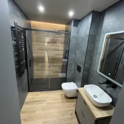 Nowoczesna łazienka z kabiną prysznicową typu walk-in, wiszącą toaletą, umywalką nablatową na szafce z szufladami, grzejnikiem drabinkowym i szarymi płytkami imitującymi marmur.