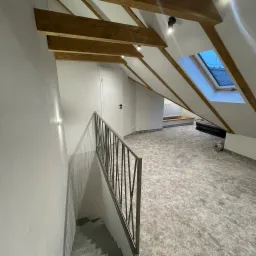 Poddasze po remoncie z drewnianymi belkami stropowymi, oknem dachowym i metalową barierką schodów, wykończone w jasnych kolorach z nową wykładziną podłogową.