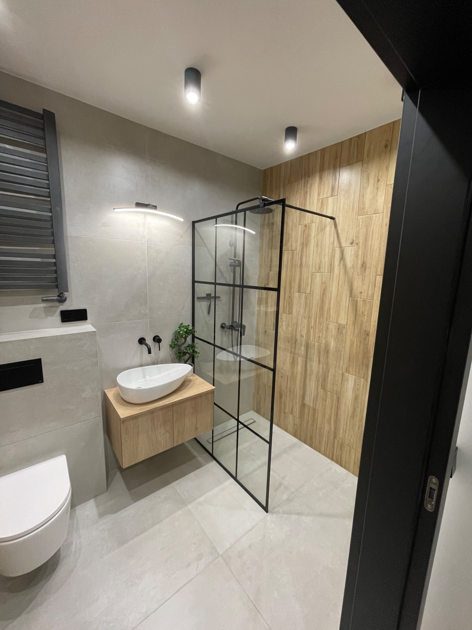 Nowoczesna łazienka z kabiną prysznicową typu walk-in z czarnymi profilami, owalną umywalką nablatową na drewnianej szafce i drewnopodobnymi płytkami na jednej ze ścian.