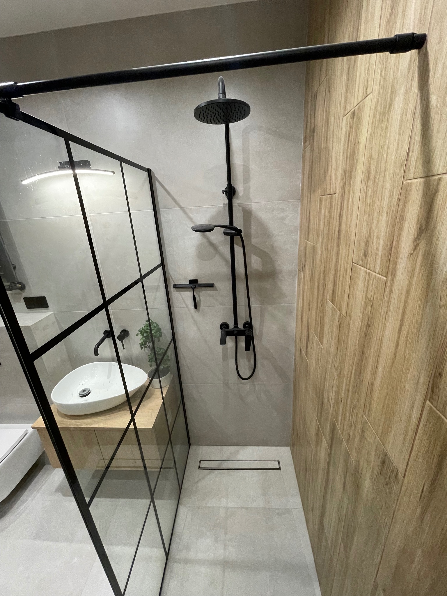 Nowoczesna łazienka z kabiną prysznicową typu walk-in z czarnymi profilami, deszczownicą i panelami ściennymi imitującymi drewno, uzupełniona białą umywalką nablatową i minimalistycznym odpływem...