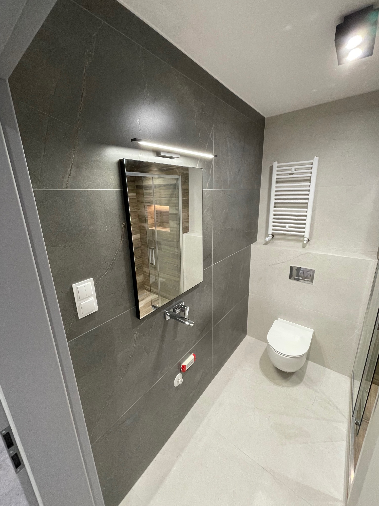 Nowoczesna łazienka z ciemnoszarą płytką imitującą kamień, podświetlanym lustrem, białym grzejnikiem drabinkowym i podwieszaną toaletą.