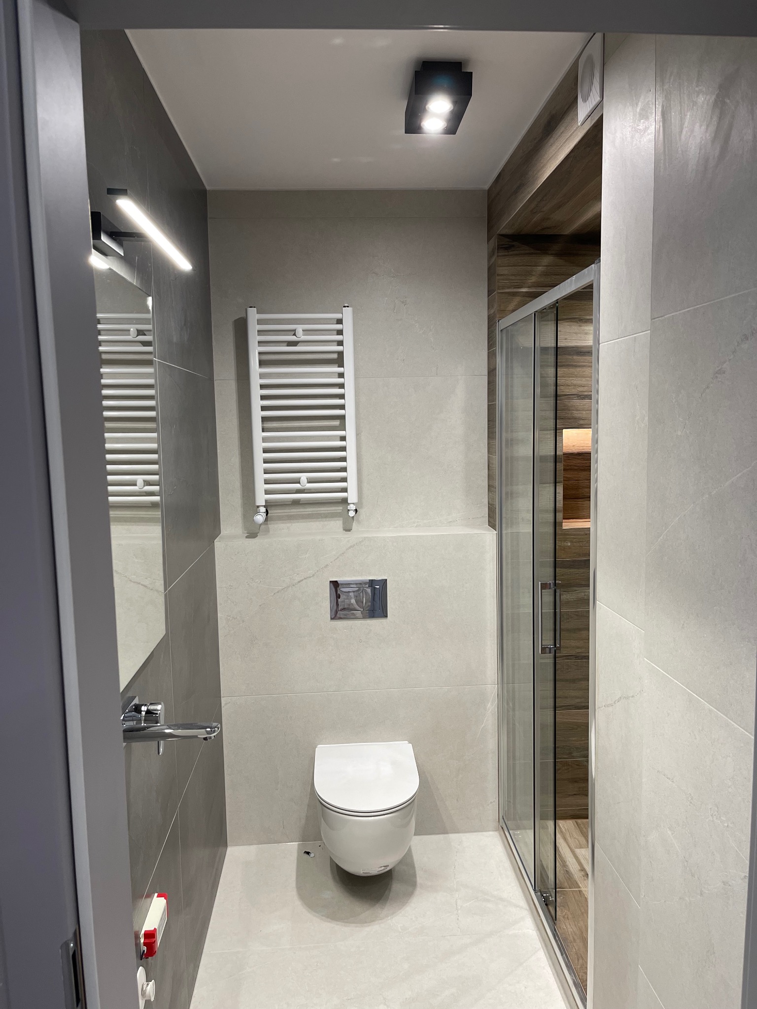 Nowoczesna łazienka z szarymi płytkami, podwieszaną toaletą, grzejnikiem drabinkowym i kabiną prysznicową z drewnianym akcentem wewnątrz.