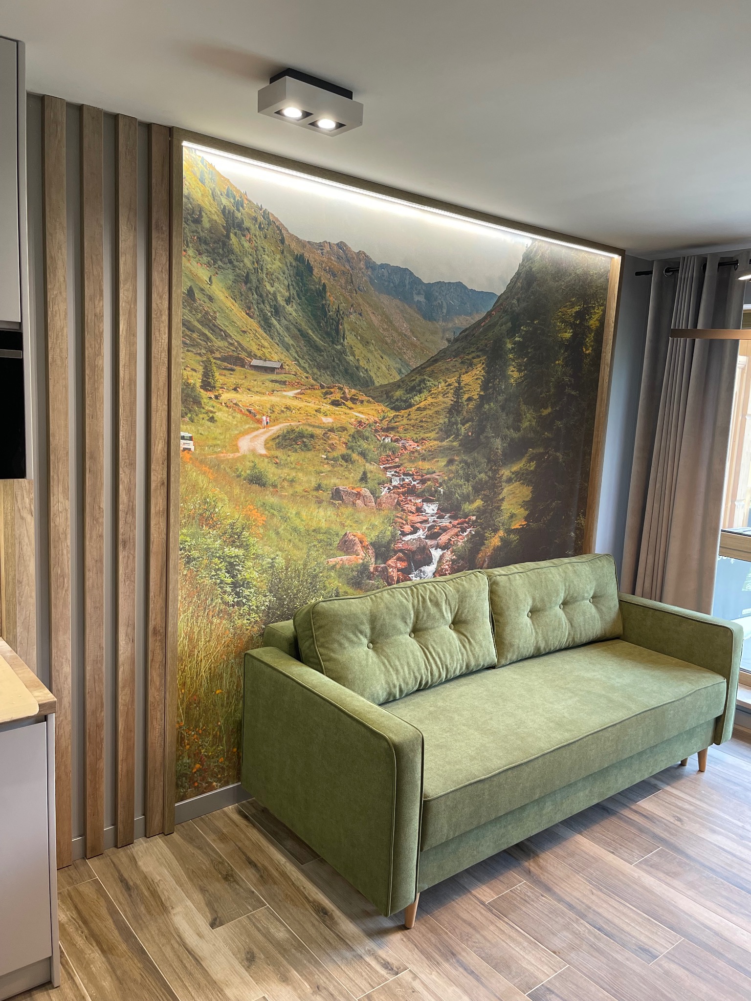 Salon z zieloną sofą na tle fototapety przedstawiającej górski krajobraz z potokiem, oświetlonej listwą LED, obok drewniane lamele na ścianie i szare zasłony przy oknie.