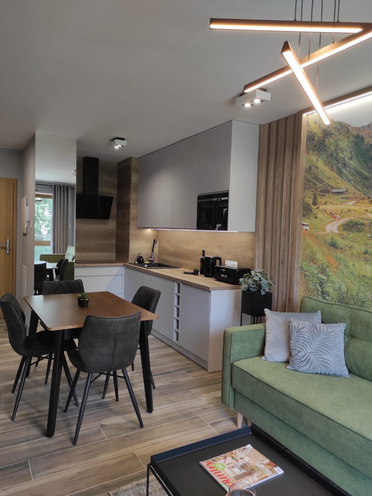 Nowoczesna kuchnia z szarymi szafkami, drewnianym blatem, stołem z czarnymi krzesłami i zieloną sofą na tle fototapety z górskim krajobrazem.