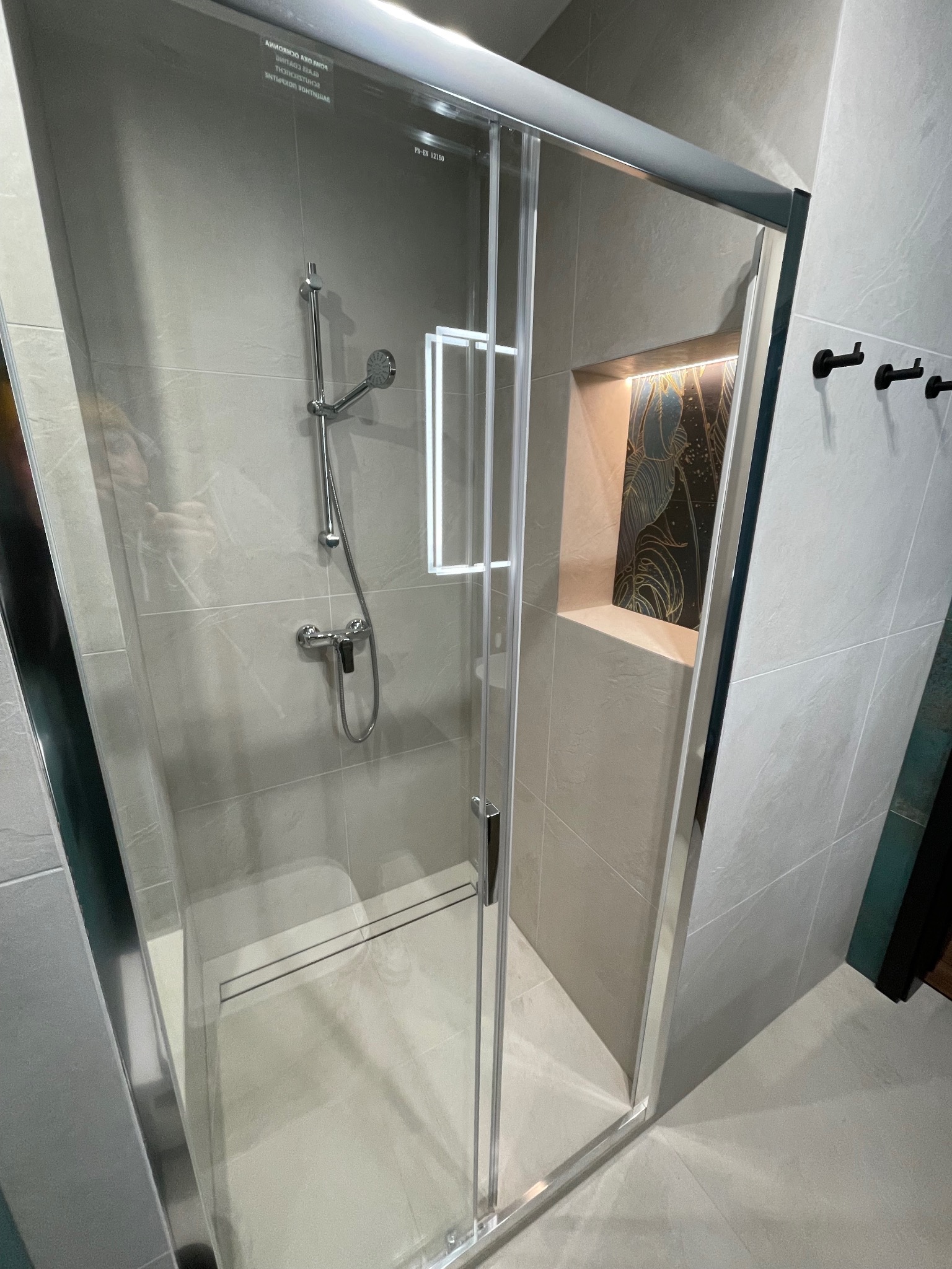 Nowoczesna kabina prysznicowa z przesuwnymi drzwiami, szare płytki imitujące kamień, wnęka z podświetleniem i dekoracyjną tapetą z motywem liści, czarne wieszaki na ścianie