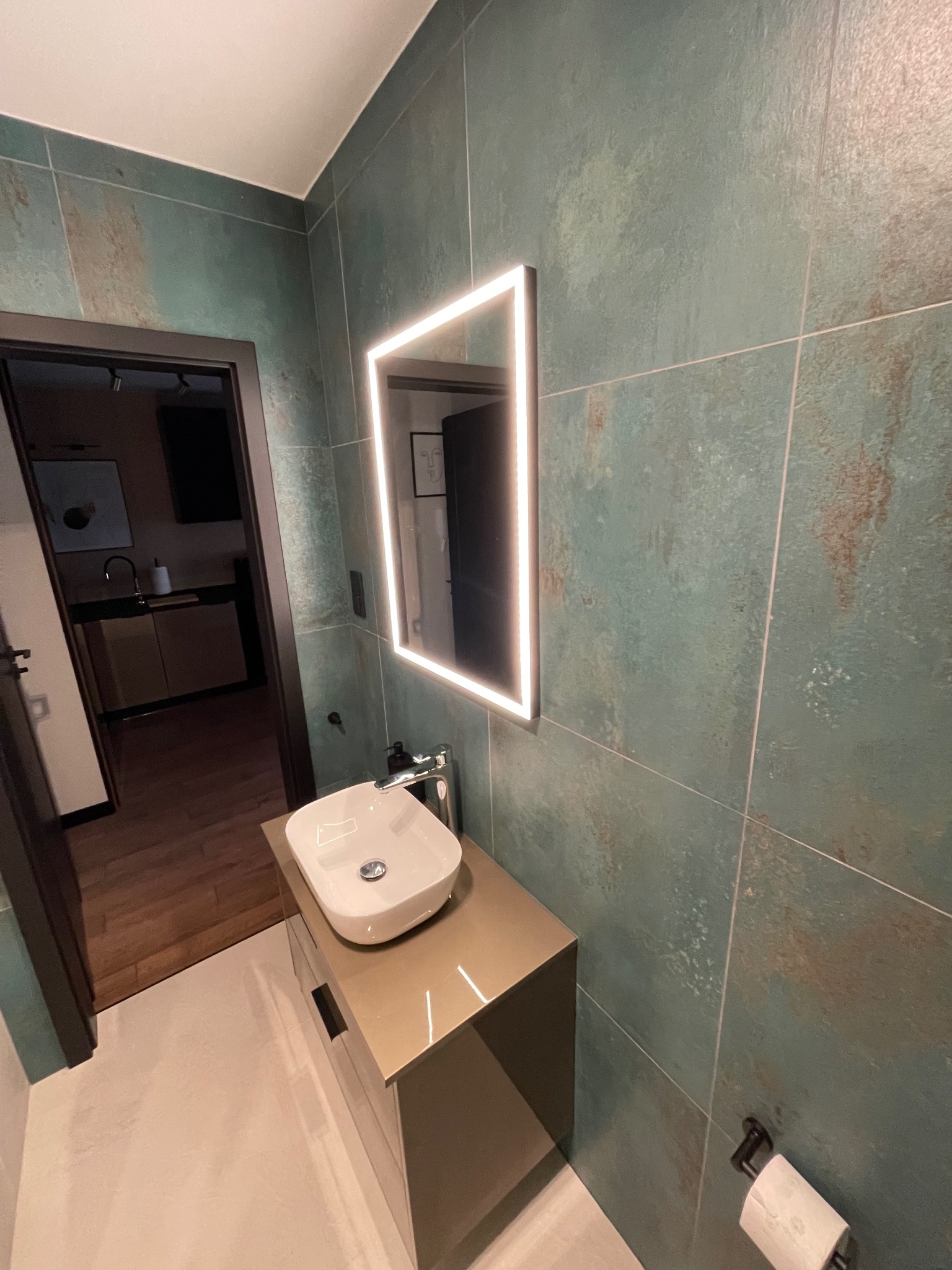 Nowoczesna łazienka z umywalką nablatową, podświetlanym lustrem w kształcie kwadratu i zielonymi płytkami imitującymi rdzę. W tle widoczne wejście do innego pomieszczenia z drewnianą podłogą.