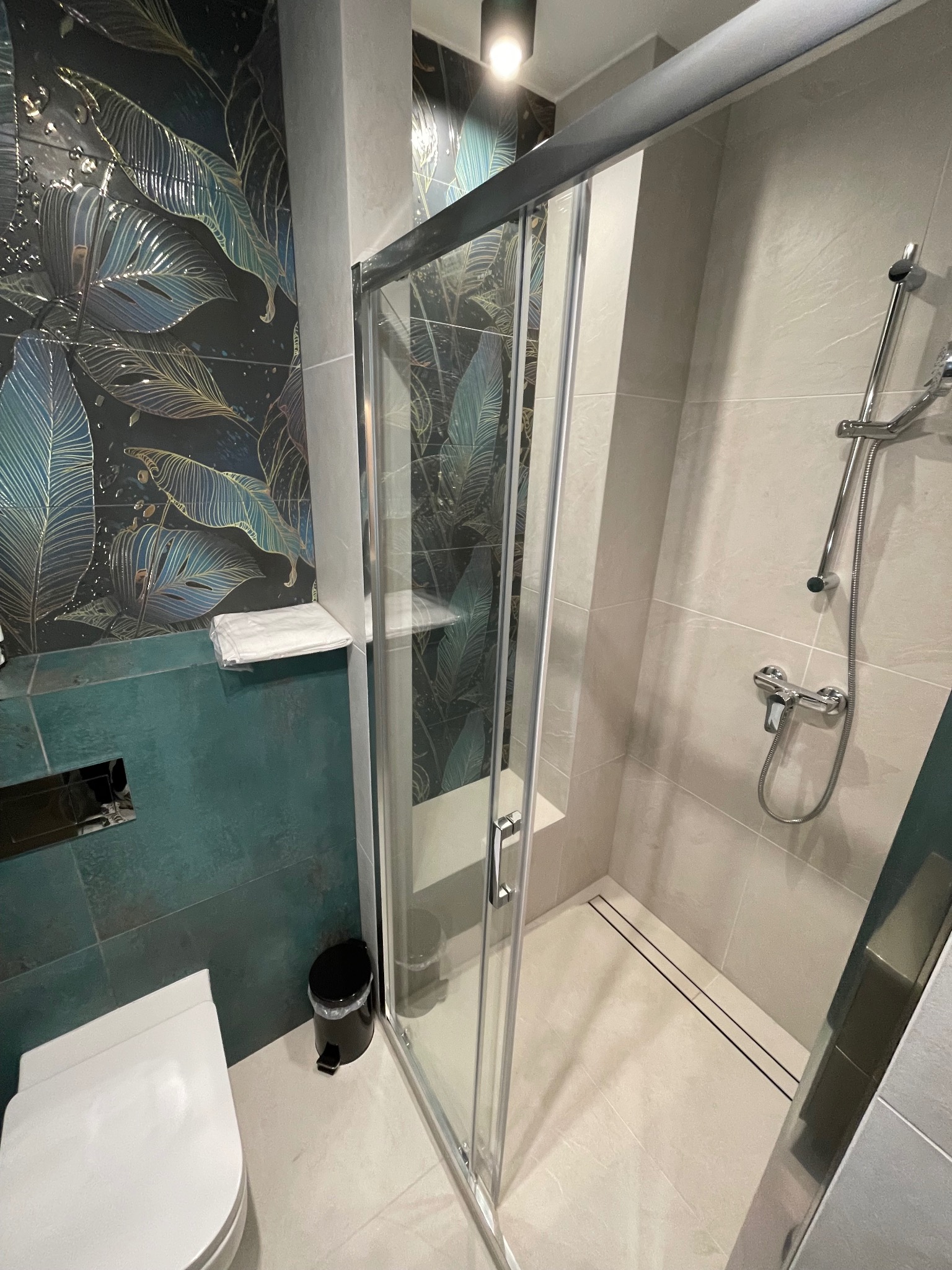 Nowoczesna łazienka z kabiną prysznicową, płytkami w motywy roślinne i minimalistyczną toaletą.