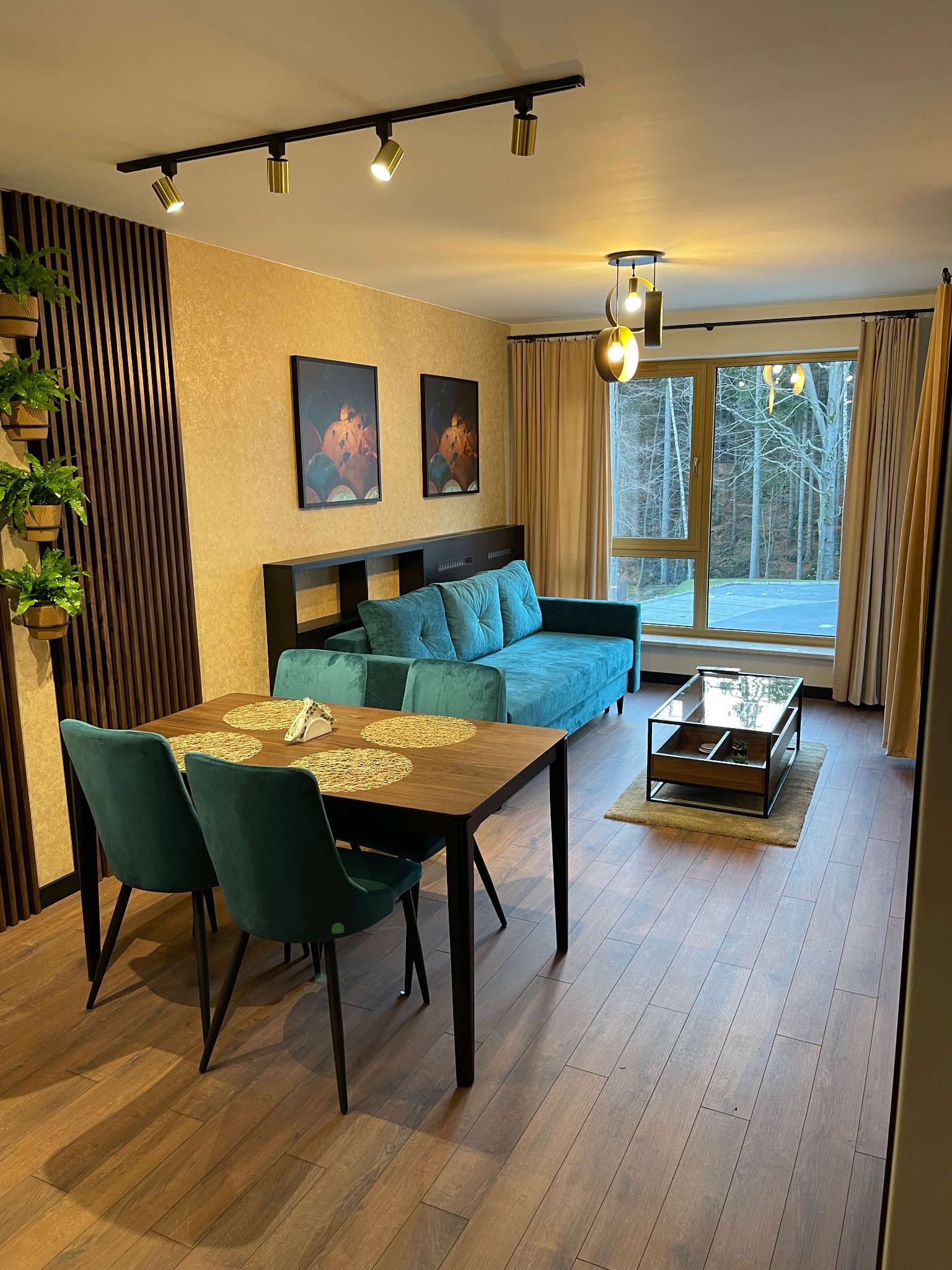 Jasne wnętrze pokoju dziennego z turkusową sofą, drewnianym stołem jadalnym i widokiem na las za oknem.