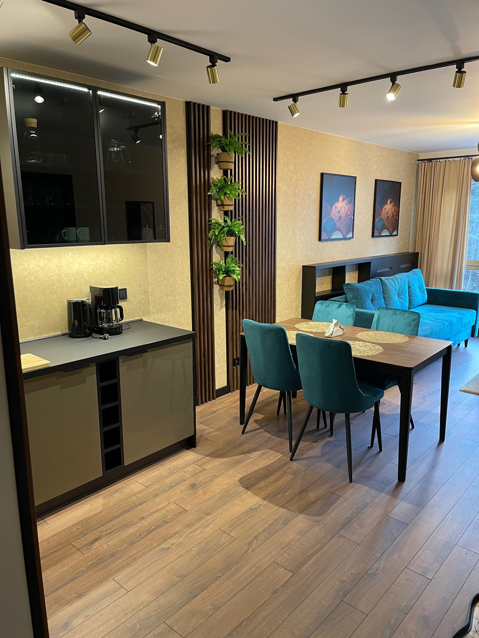 Jasne wnętrze mieszkania z drewnianą podłogą, szafkami kuchennymi z czarnymi akcentami, stołem z turkusowymi krzesłami, turkusową sofą i pionowym ogrodem z roślinami doniczkowymi