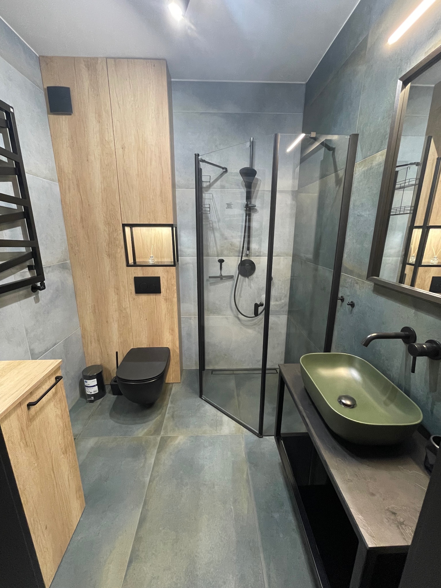 Nowoczesna łazienka z kabiną prysznicową typu walk-in, umywalką w odcieniu oliwkowej zieleni, czarną armaturą i toaletą w tym samym kolorze, drewniane szafki z akcentami czerni oraz szare płytki...