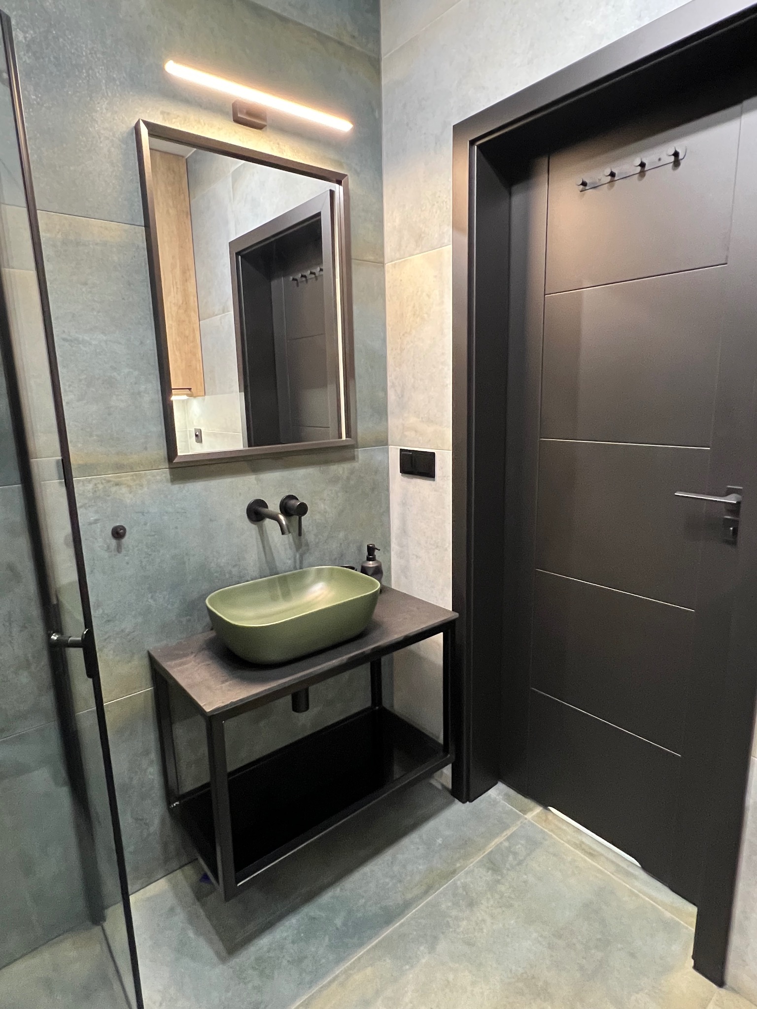 Nowoczesna łazienka z zieloną umywalką nablatową, czarną armaturą i ramą lustra, oświetleniem LED oraz ciemnymi płytkami na ścianach i podłodze, obok ciemne drzwi z wieszakami.