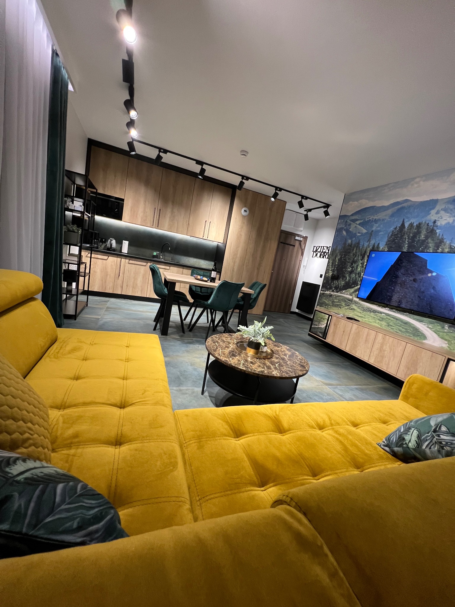 Nowoczesne wnętrze mieszkania z żółtą sofą, drewnianą zabudową kuchenną, stołem z krzesłami i telewizorem z fototapetą górską.