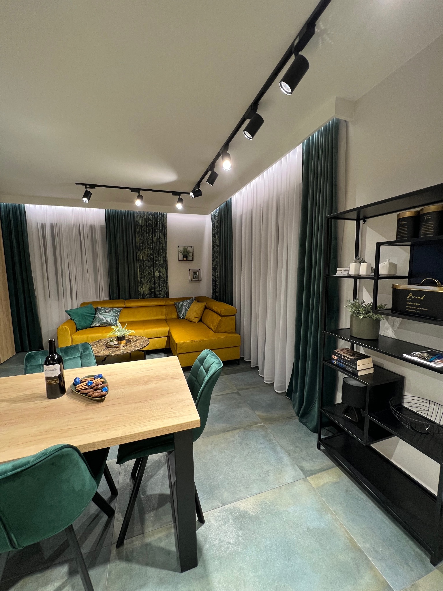Nowoczesne wnętrze z żółtą sofą narożną, zielonymi zasłonami i krzesłami, drewnianym stołem z przekąskami i butelką wina, oświetleniem punktowym na suficie i czarnym regałem.