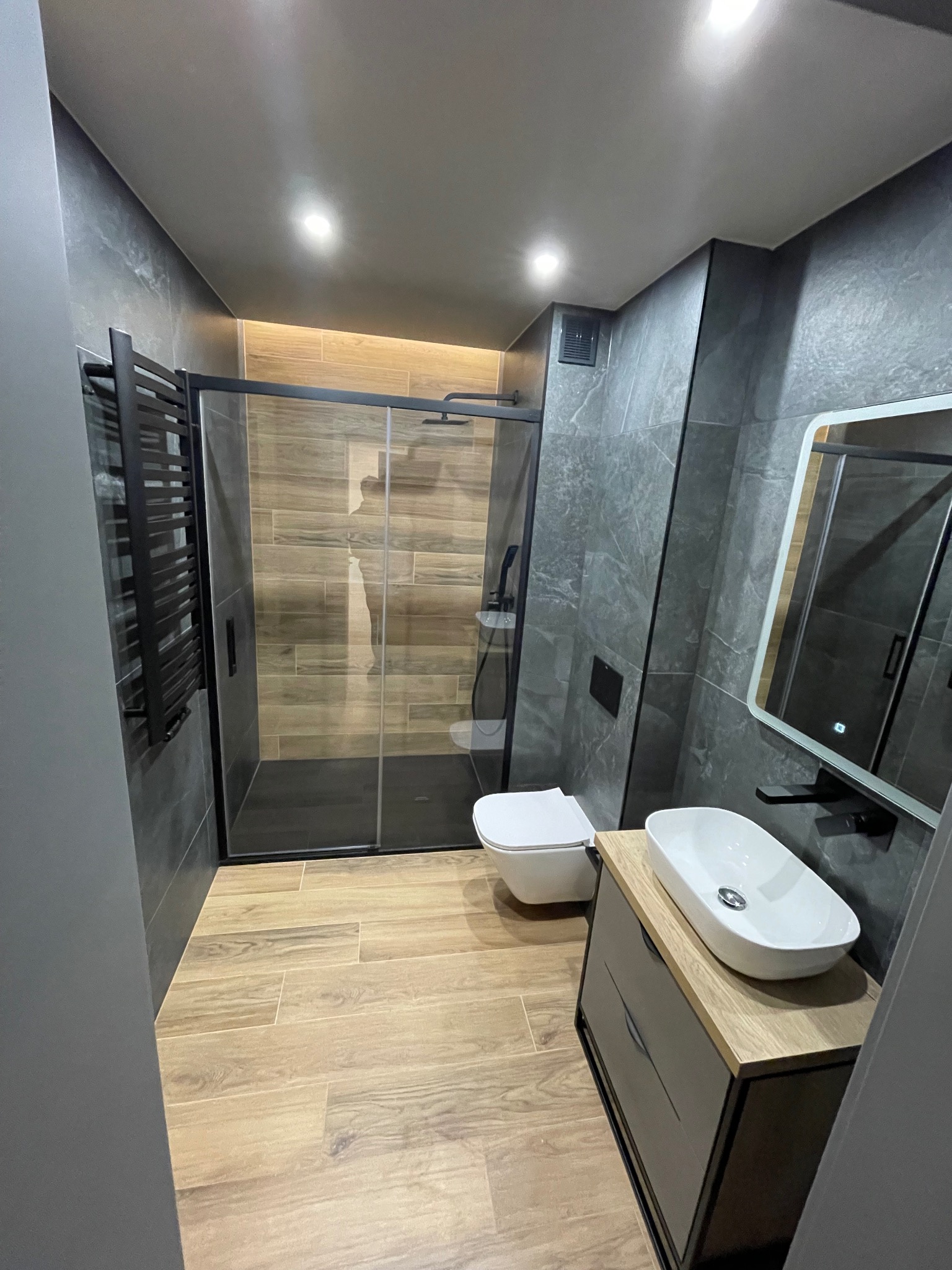 Nowoczesna łazienka z kabiną prysznicową typu walk-in, wiszącą toaletą, umywalką nablatową na szafce z szufladami, grzejnikiem drabinkowym i szarymi płytkami imitującymi marmur.