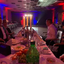 Długi st&oacute;ł udekorowany świecami w świecznikach, owocami i naczyniami, oświetlony czerwono-niebieskimi światłami, z widoczną sceną w tle i siedzącymi gośćmi.