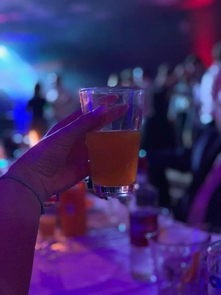 Podświetlony drink w szklance trzymany w dłoni na imprezie w Olszewce, w tle rozmazane sylwetki osób i stoliki z napojami.