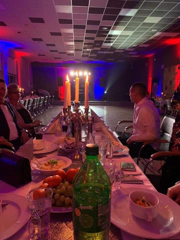 Długi stół udekorowany świecami w świecznikach, owocami i naczyniami, oświetlony czerwono-niebieskimi światłami, z widoczną sceną w tle i siedzącymi gośćmi.