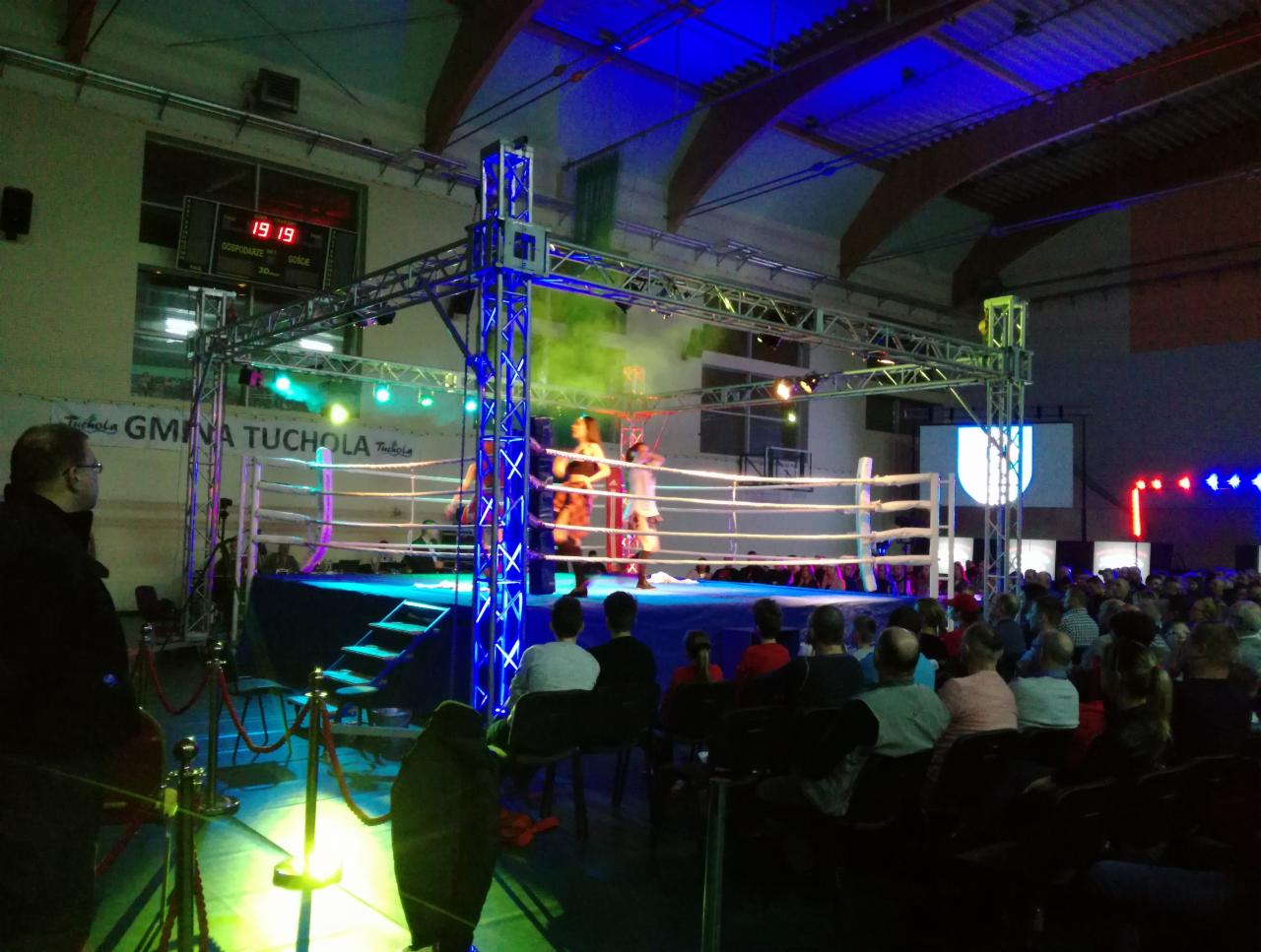Oświetlony ring bokserski z publicznością, konstrukcja kratowa z niebieskim oświetleniem, zielony dym, tablica wyników i logo Gminy Tuchola w tle.